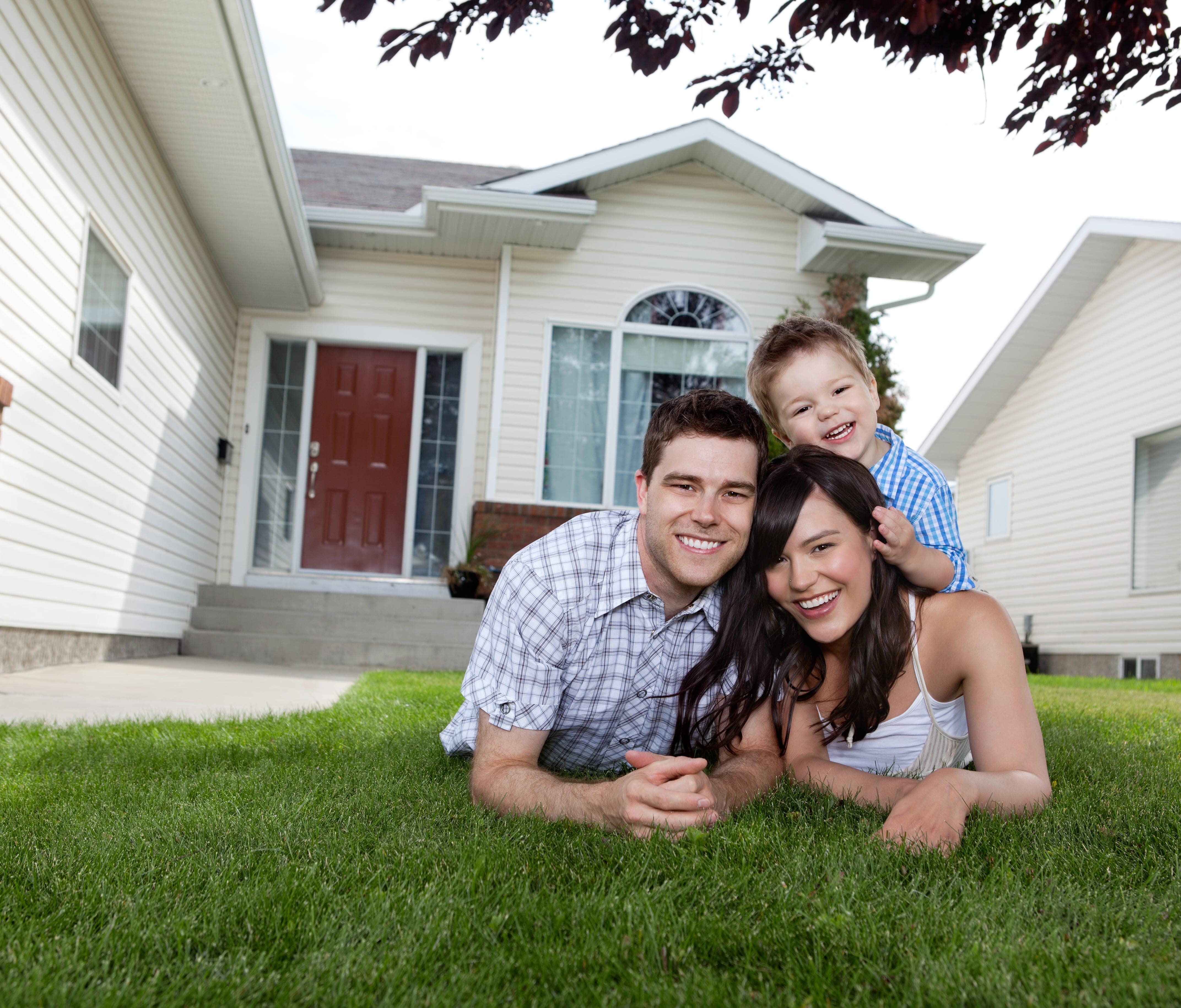Man, Woman, and Child Smile While Laying on the Grass Lawn in Front of a House