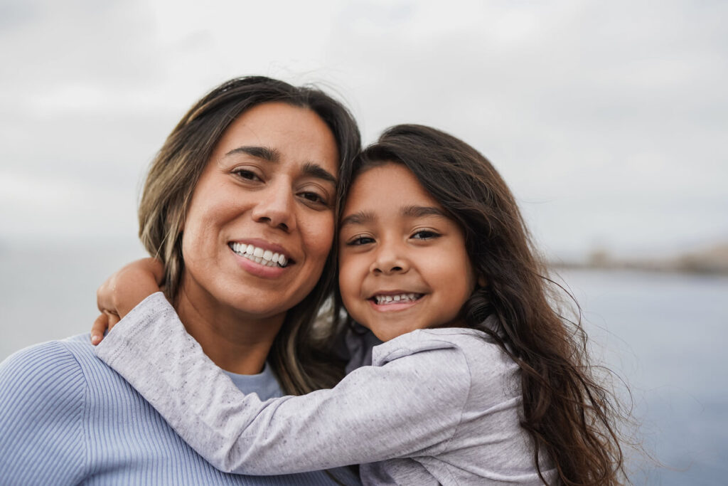 Latina mother and young daughter smiling and hugging by the ocean in San Diego.