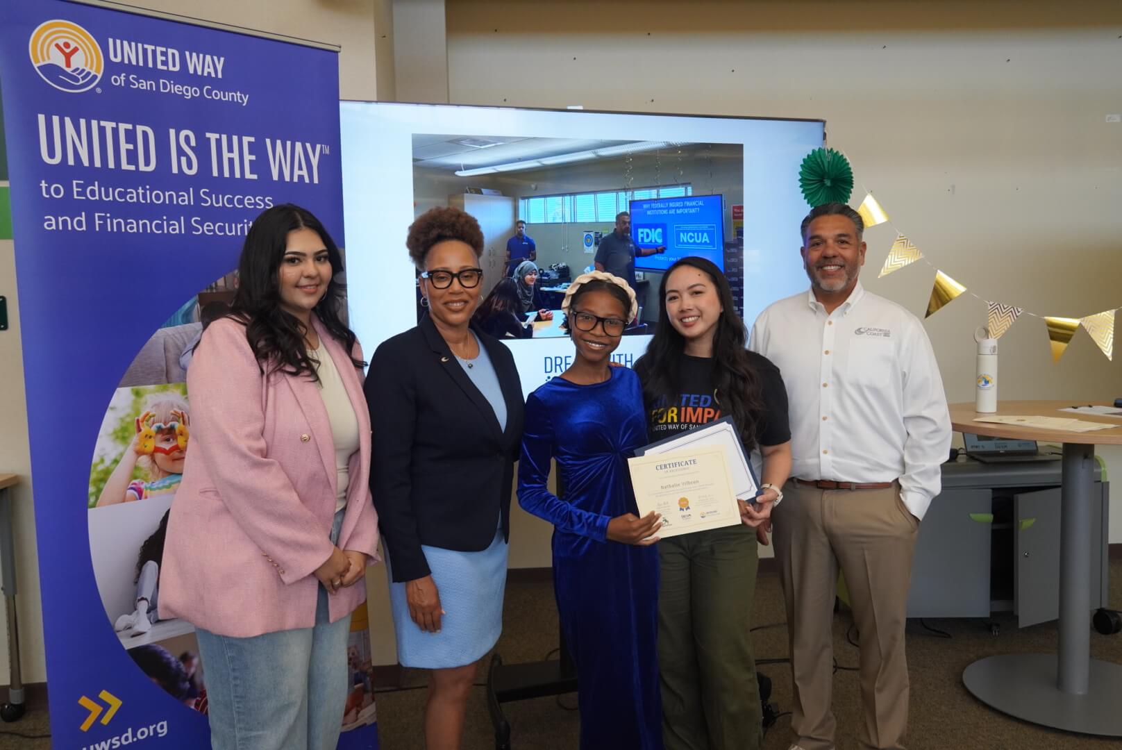 Dream with STEAM student proudly holding a certificate with two members of Cal Coast Credit Union and United Way of San Diego County with a screen in the background showing a photo from a financial literacy course and a banner that says "United Is The Way"