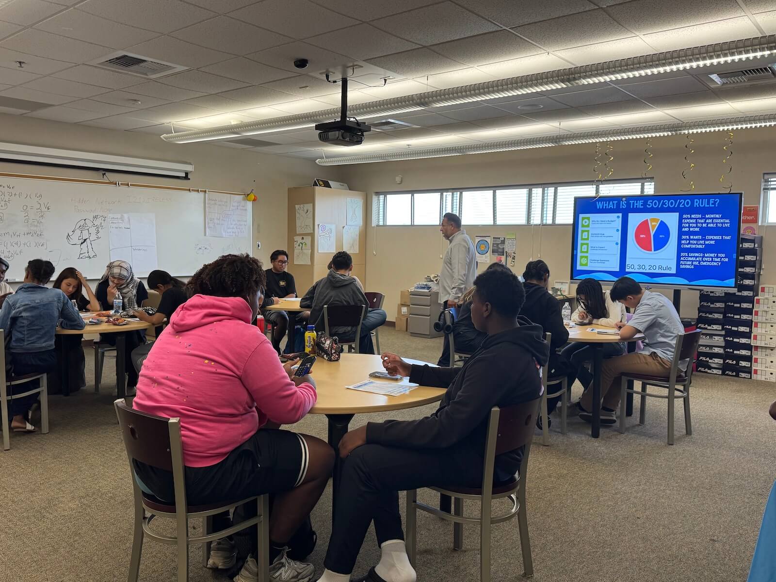 A classroom with Dream with STEAM students being instructed by someone from Cal Coast Credit Union on Financial Literacy with a screen displaying information on the 50/30/20 rule.